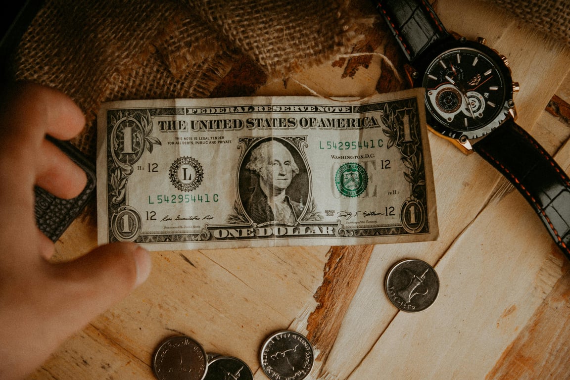 A person reaches for a one dollar bill surrounded by coins and a watch on a wooden table.