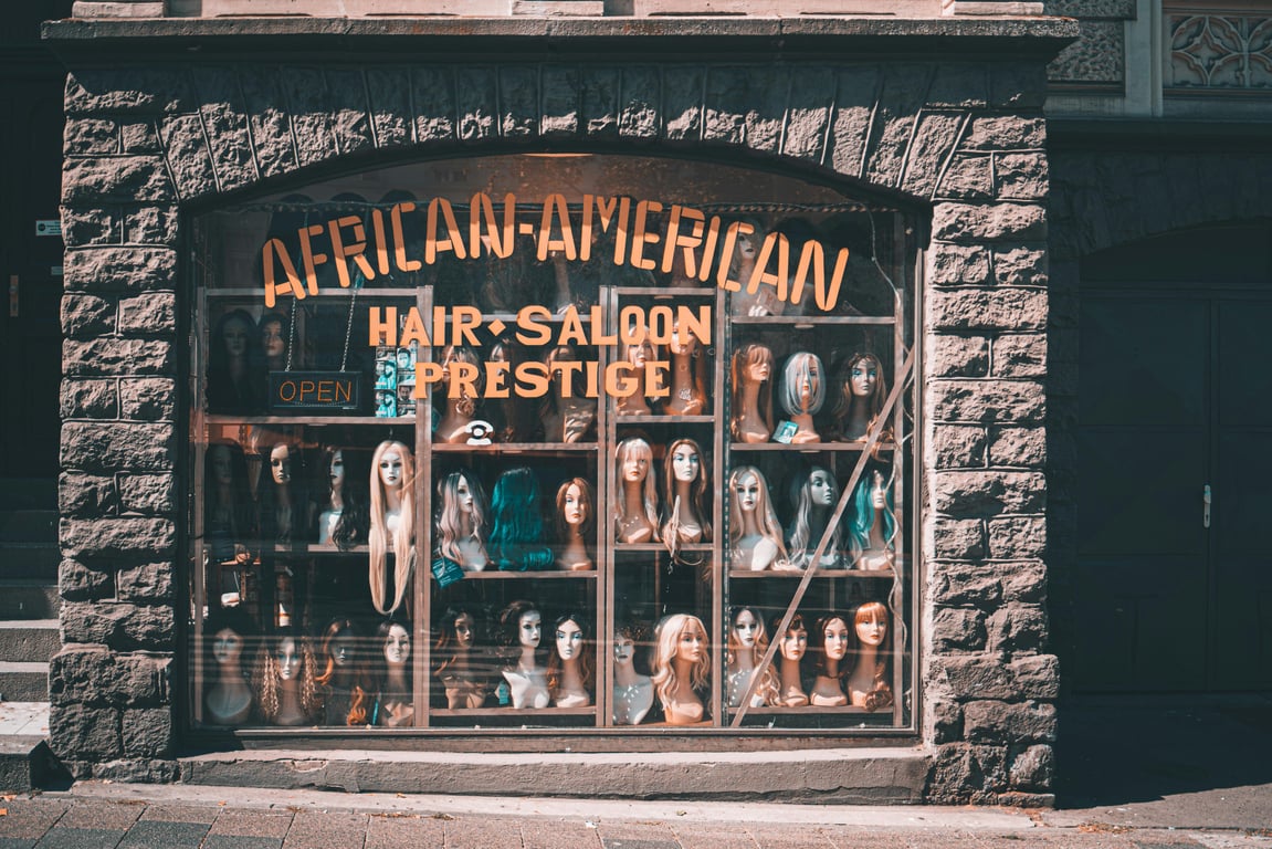 Showcase of various wigs in a store window labeled 'African-American Hair Saloon Prestige'.