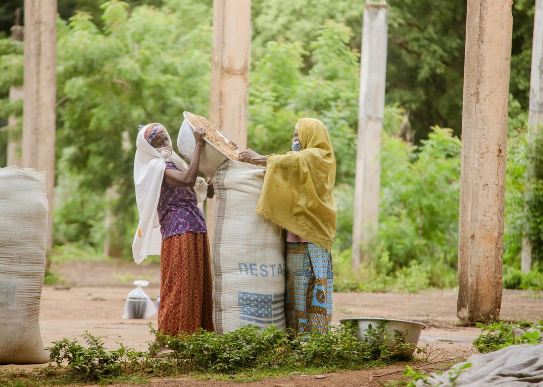 Two women in traditional attire working with large sacks in a Ghanaian outdoor environment.