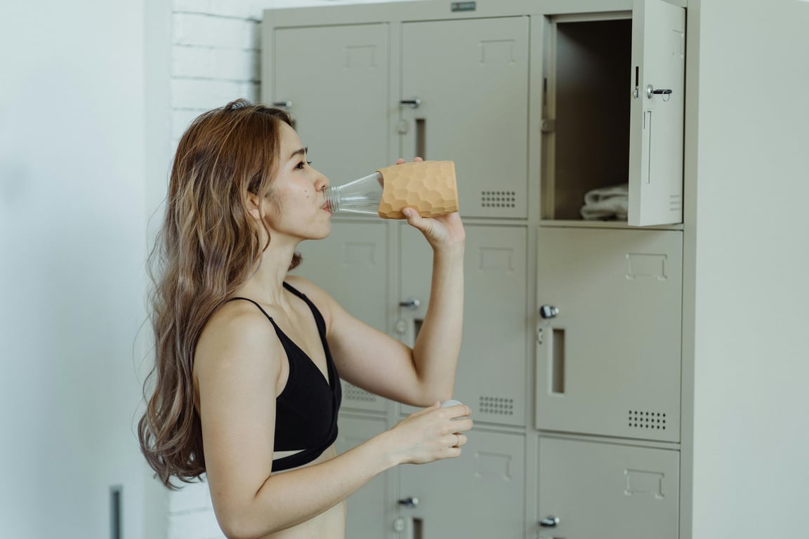 Young woman in sportswear drinking water from a glass in a locker room, emphasizing hydration and fitness.