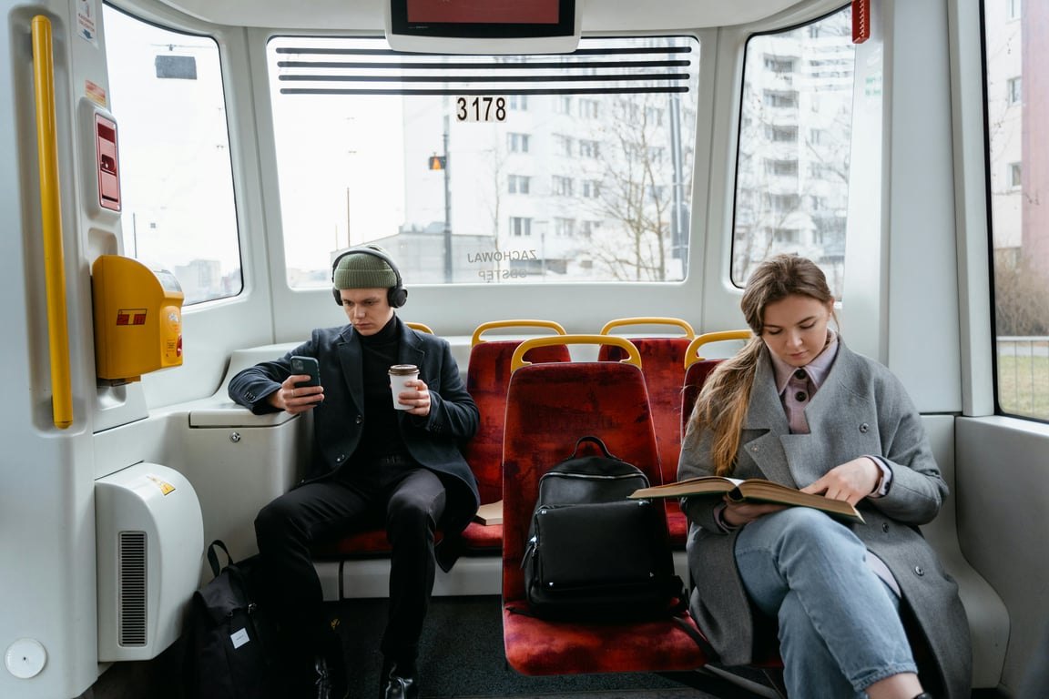 Two commuters on a tram, using gadgets and reading, showcasing a typical urban lifestyle.