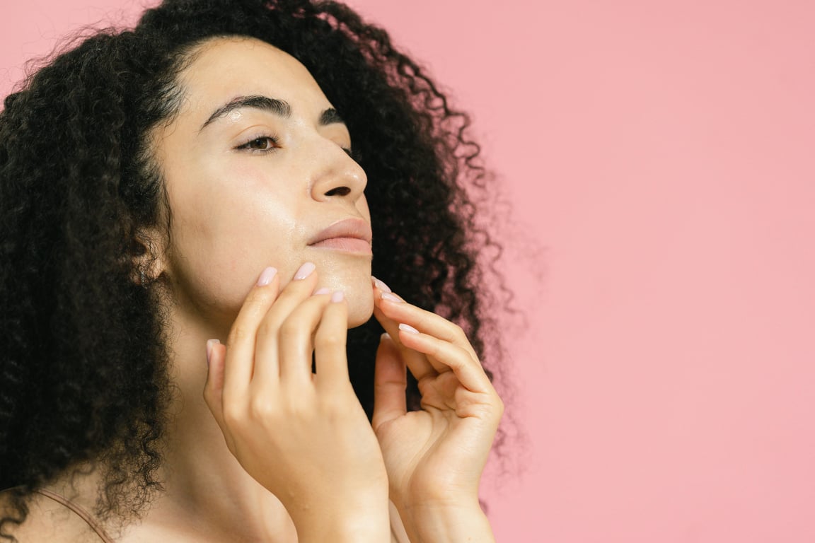 A woman with curly hair touches her face, set against a pastel pink background.