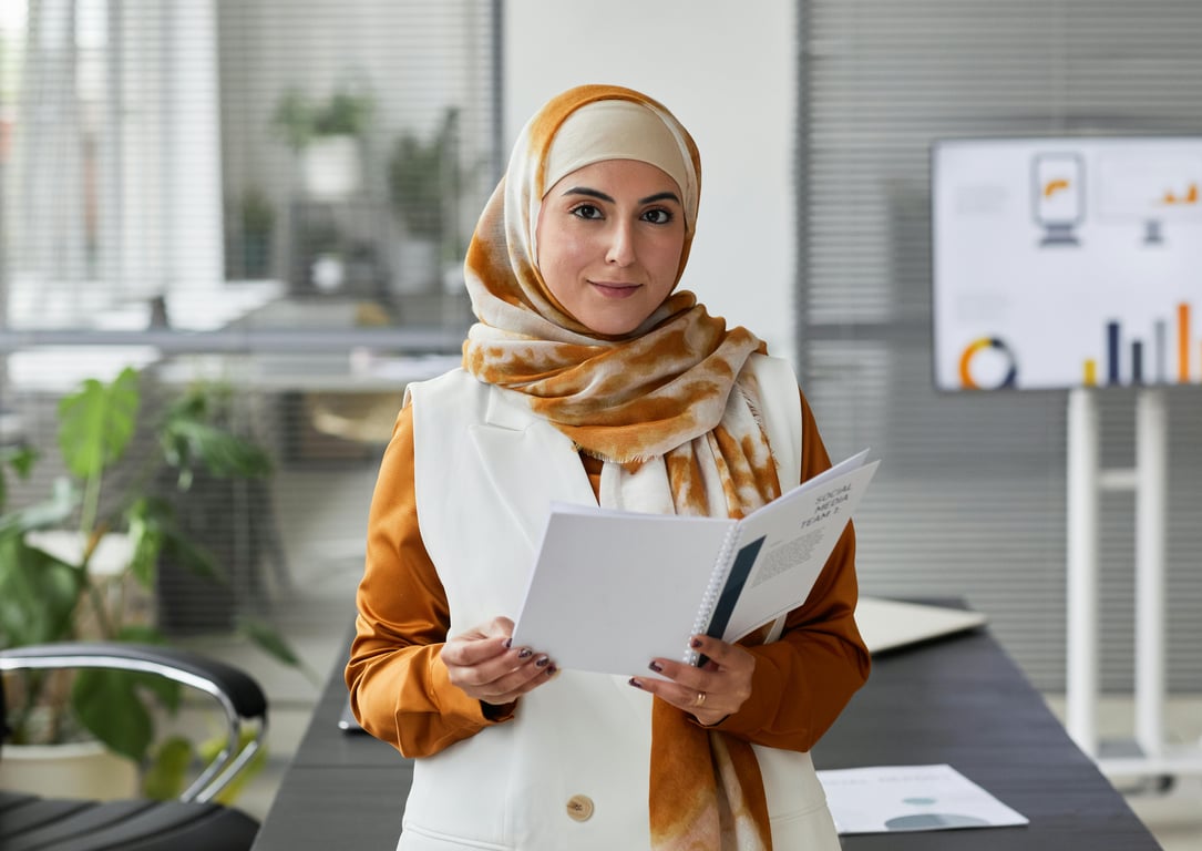 A professional Muslim woman in an office setting, confidently holding documents and smiling.