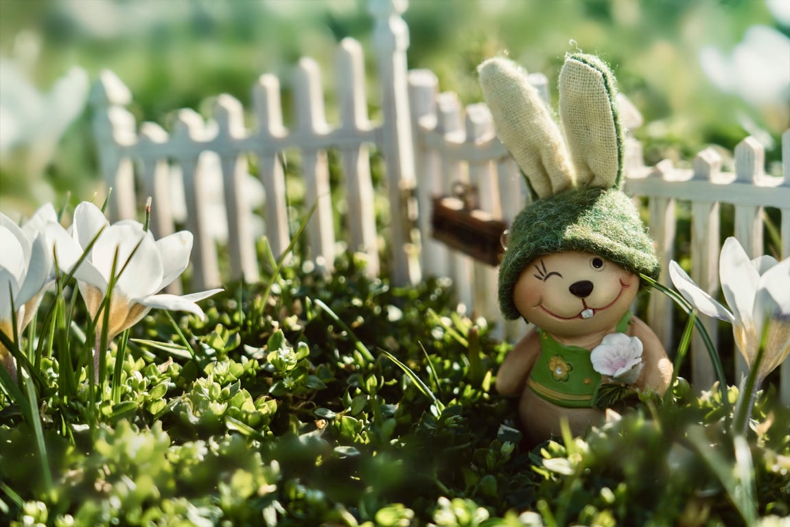 Adorable bunny figurine in a sunny garden surrounded by blooming flowers and a white picket fence.