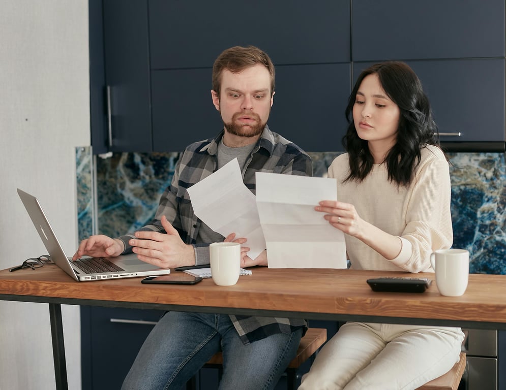 A couple sitting at a wooden table, reviewing documents with a laptop nearby, expressing concern.