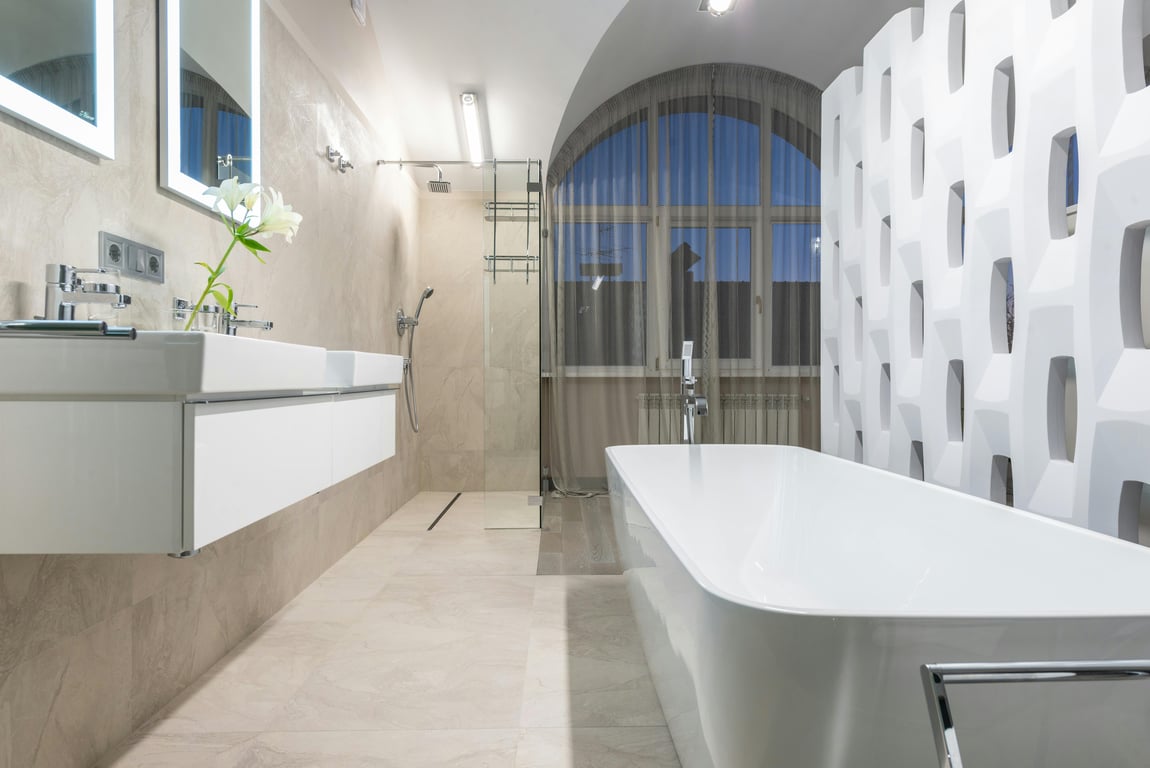 White bathtub near sink with flowers and mirror placed in light modern bathroom with glowing lamp and window in apartment