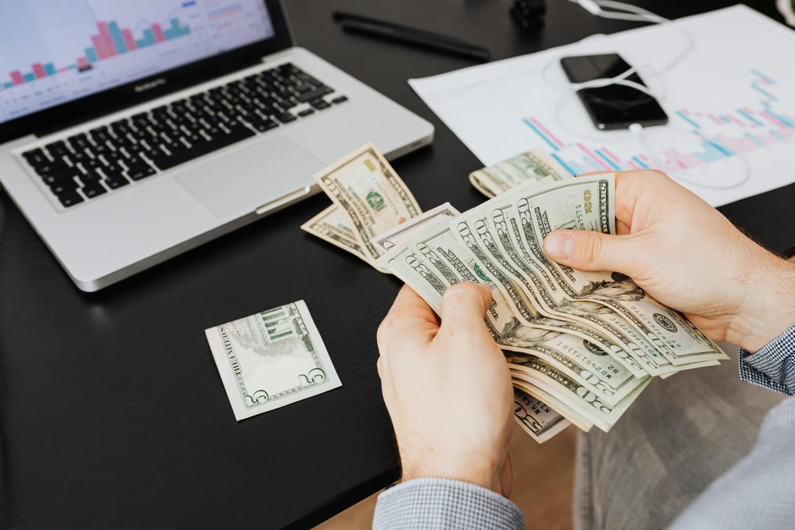 Person counting dollar bills at desk with laptop and financial charts, symbolizing finance management.