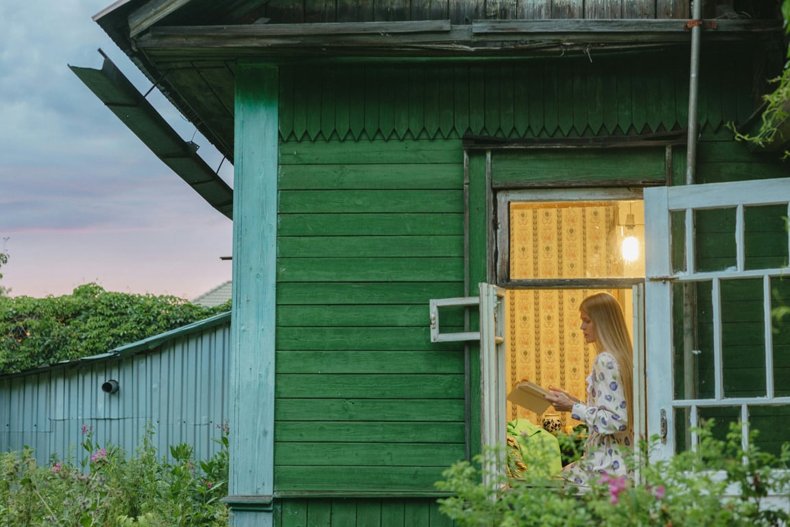 A serene scene of a woman reading inside a wooden house at sunset.