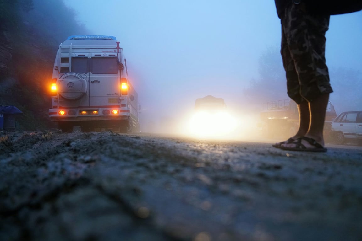 A foggy morning on a road with vehicles and a traveler, headlights piercing through the mist.