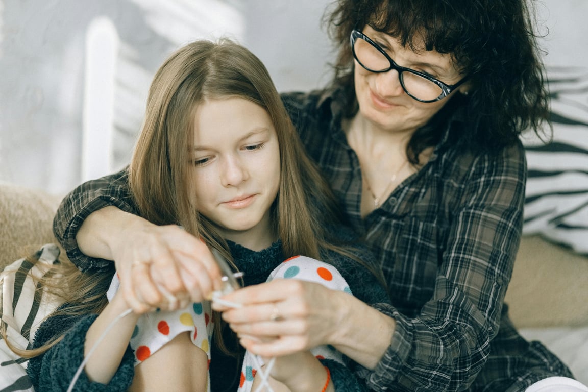 A mother guides her daughter in knitting, fostering a bond through learning at home.