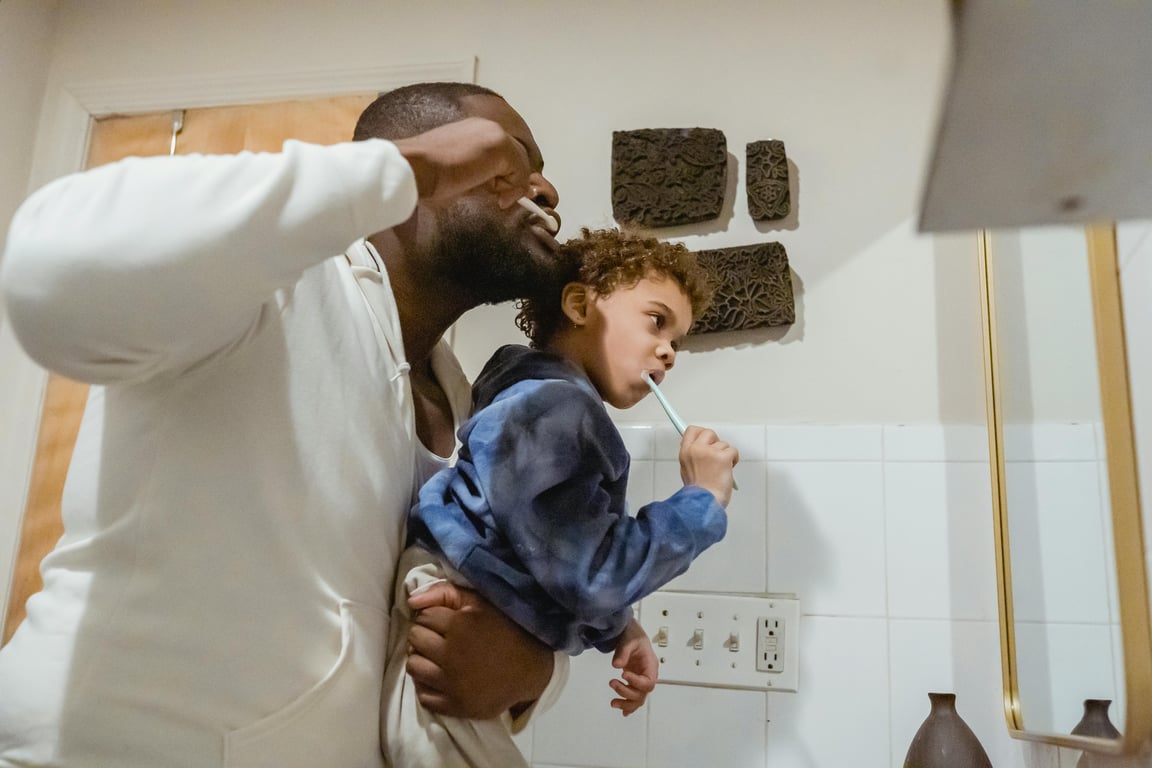 Side view of focused African American father with boy in hands brushing teeth with toothbrushes during morning routine in bathroom