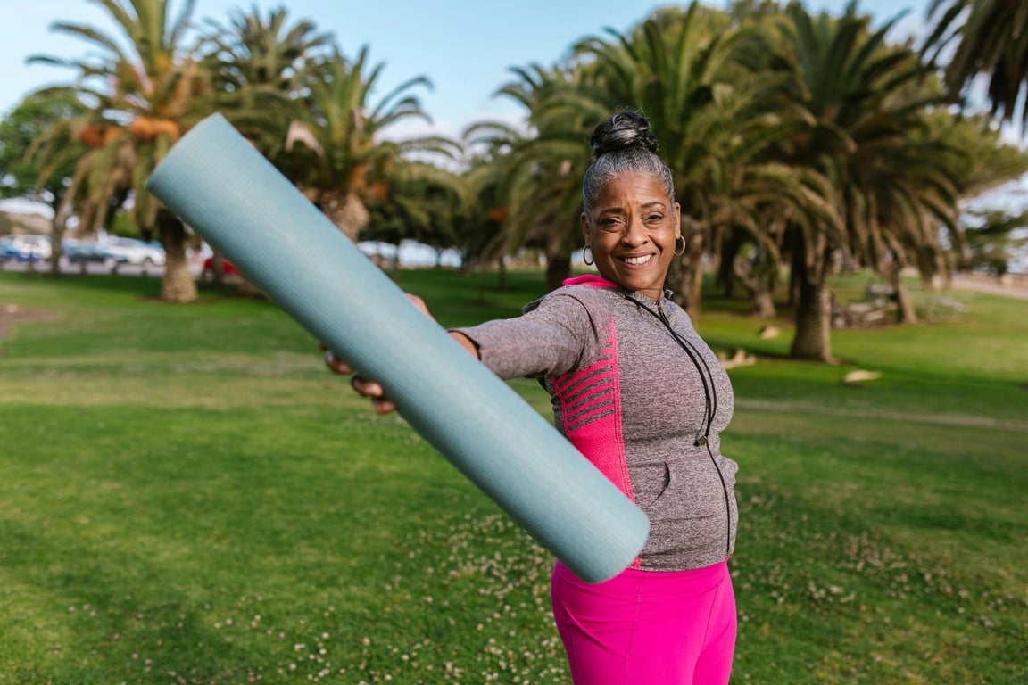 Smiling senior woman holding yoga mat, preparing for outdoor exercise in a sunny park.
