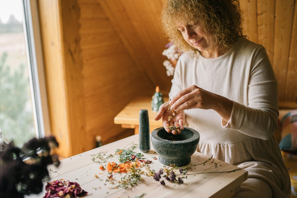 A woman in a white dress using a mortar and pestle with dried herbs on a wooden table.