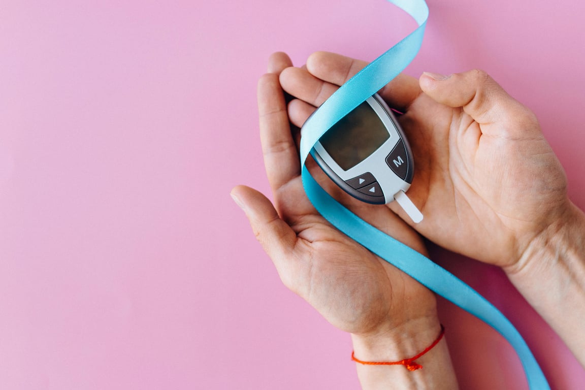 Hands holding a glucometer with a blue ribbon symbolizing diabetes awareness on a pink background.