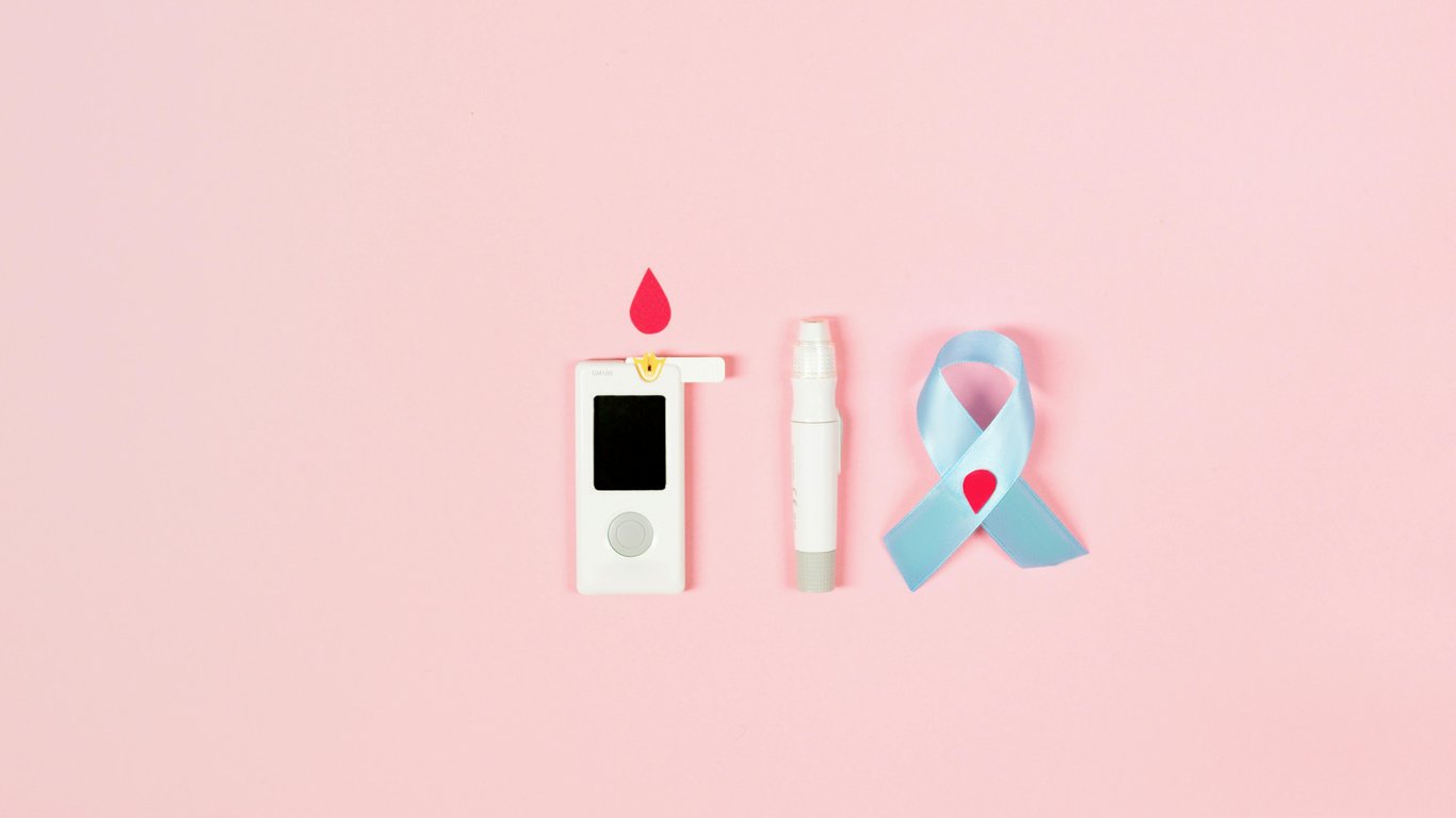 Flat lay of a glucometer, lancet pen, and diabetes awareness ribbon on a pink background symbolizing health awareness.