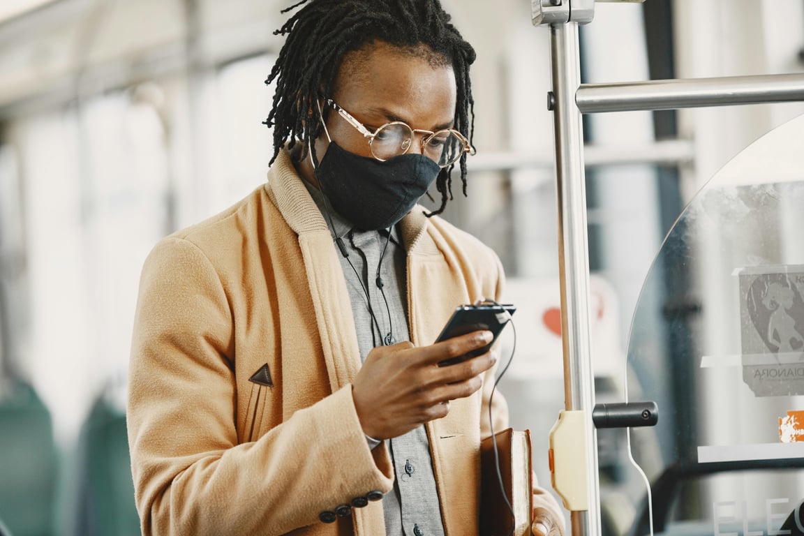 Stylish man with dreadlocks wearing a face mask on public transportation, checking his phone. Urban lifestyle context.