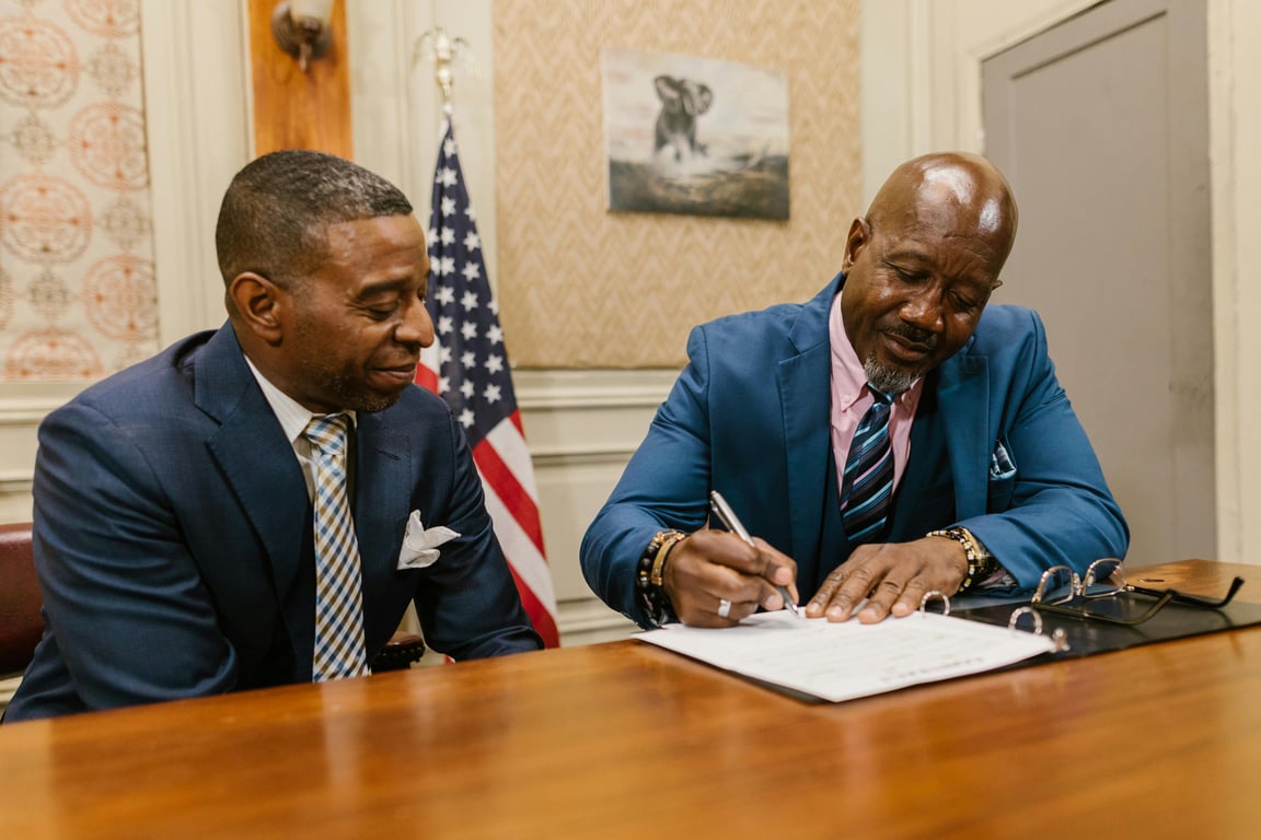 Two businessmen in suits signing documents in an office setting with an American flag.