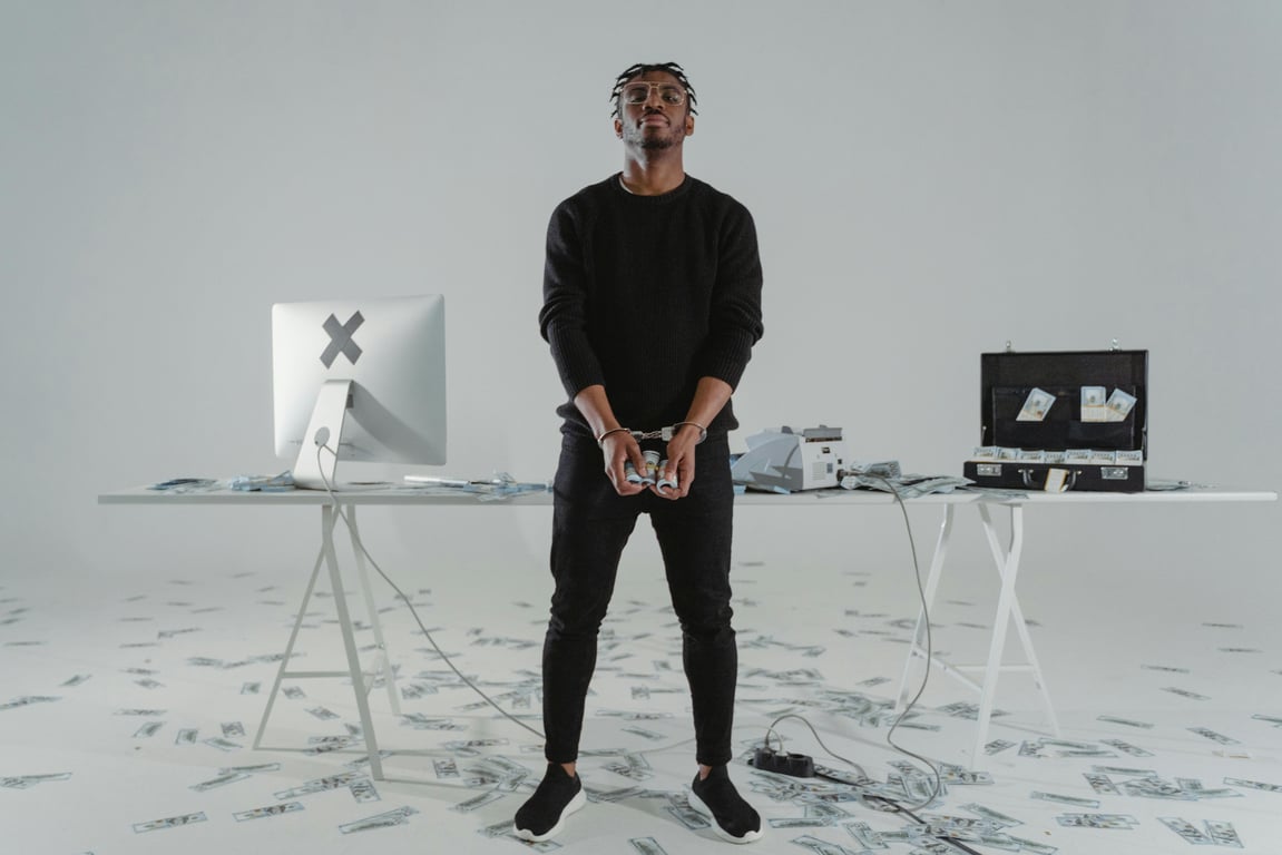 A handcuffed man stands in a room with scattered money, a computer, and a briefcase.