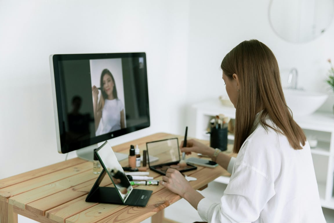 A woman follows an online makeup tutorial using a computer and tablet at home.