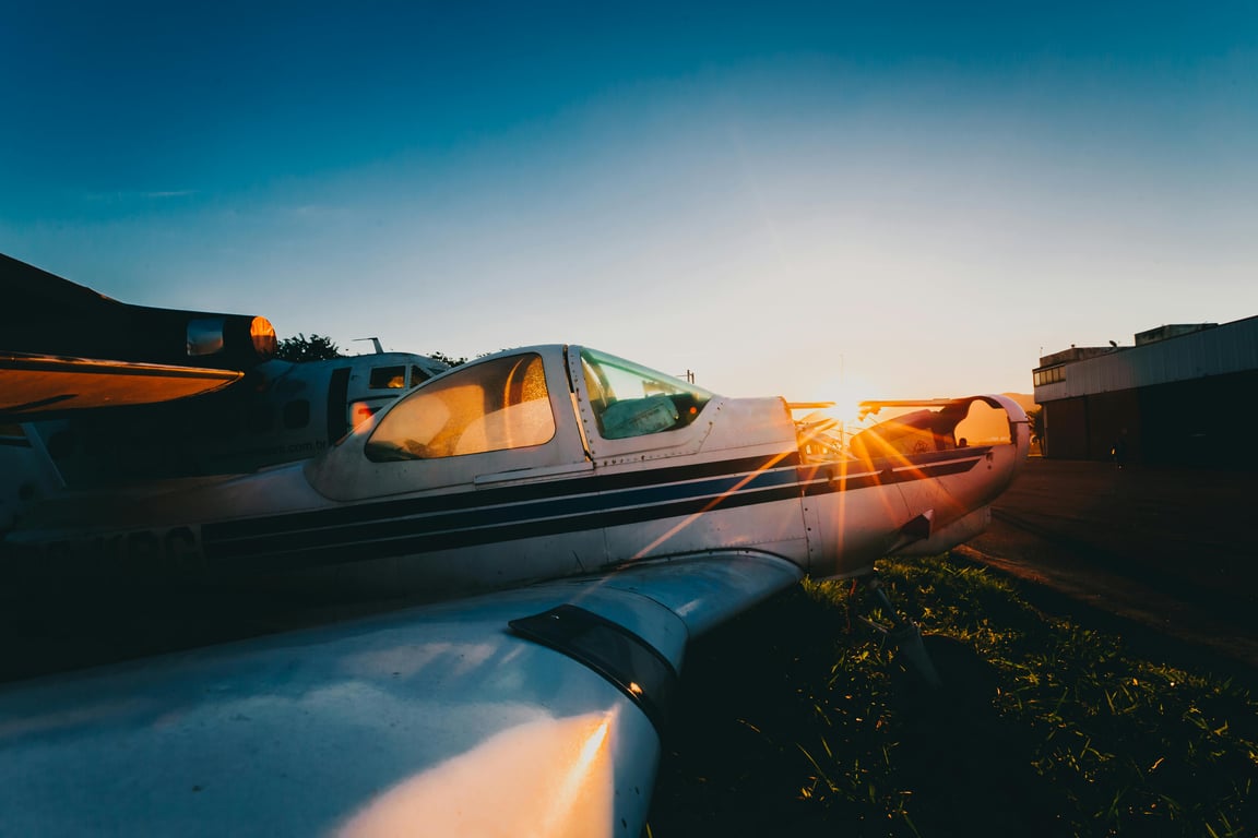 A small aircraft parked on the runway with sunset light creating a dramatic sun glare on its wing.