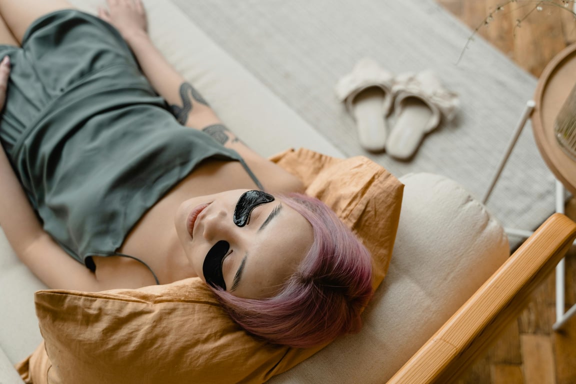 A woman with pink hair relaxes on a couch wearing under eye masks for skincare.