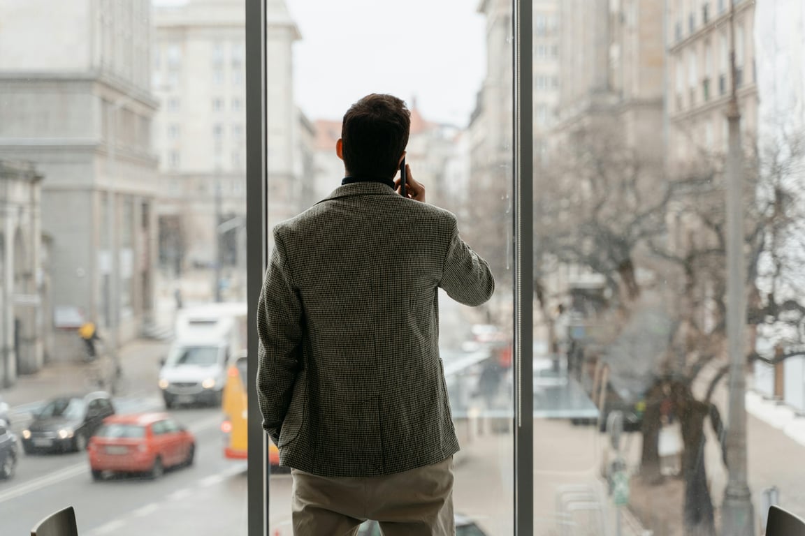 A businessman on a phone call, standing next to a window overlooking a busy city street.