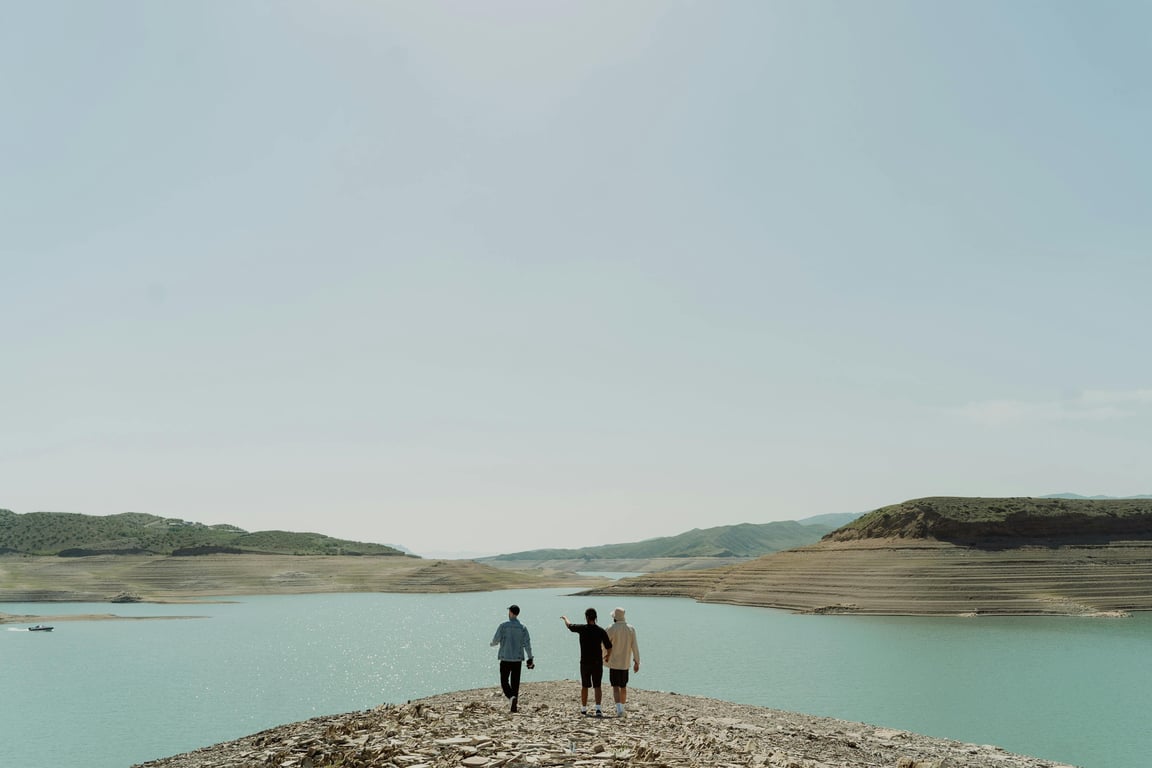 Three people walking by a serene lake under a clear sky, surrounded by hills.