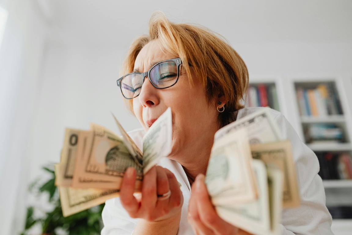 A happy woman with eyeglasses cherishing US dollar bills indoors, symbolizing wealth and financial success.