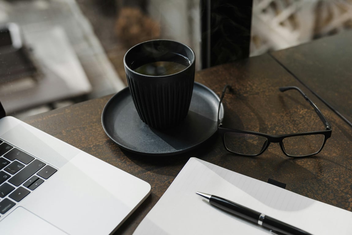 A sleek office desk setup featuring coffee, a laptop, glasses, pen, and notebook.