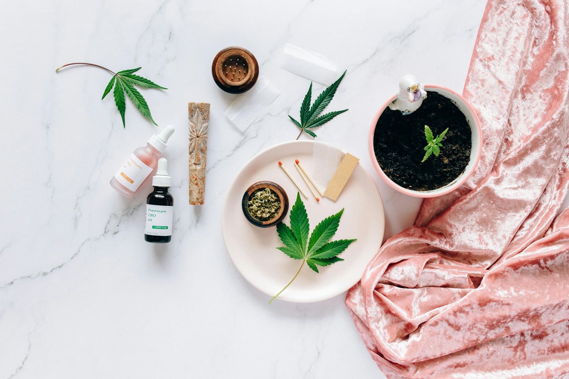 Flat lay of cannabis leaves, CBD oils, and accessories on marble surface.