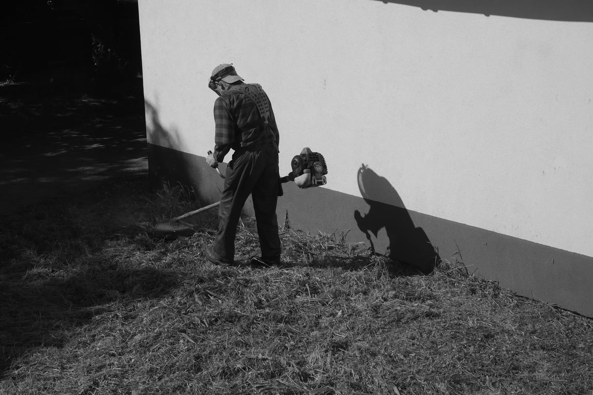 Black and white photo of a person trimming grass with a weed whacker next to a wall.
