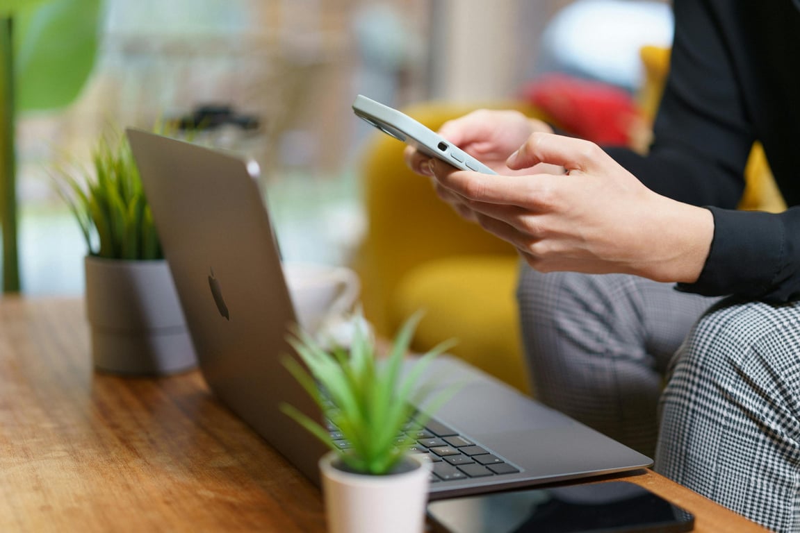 A person working on a smartphone and laptop at home with plants nearby.