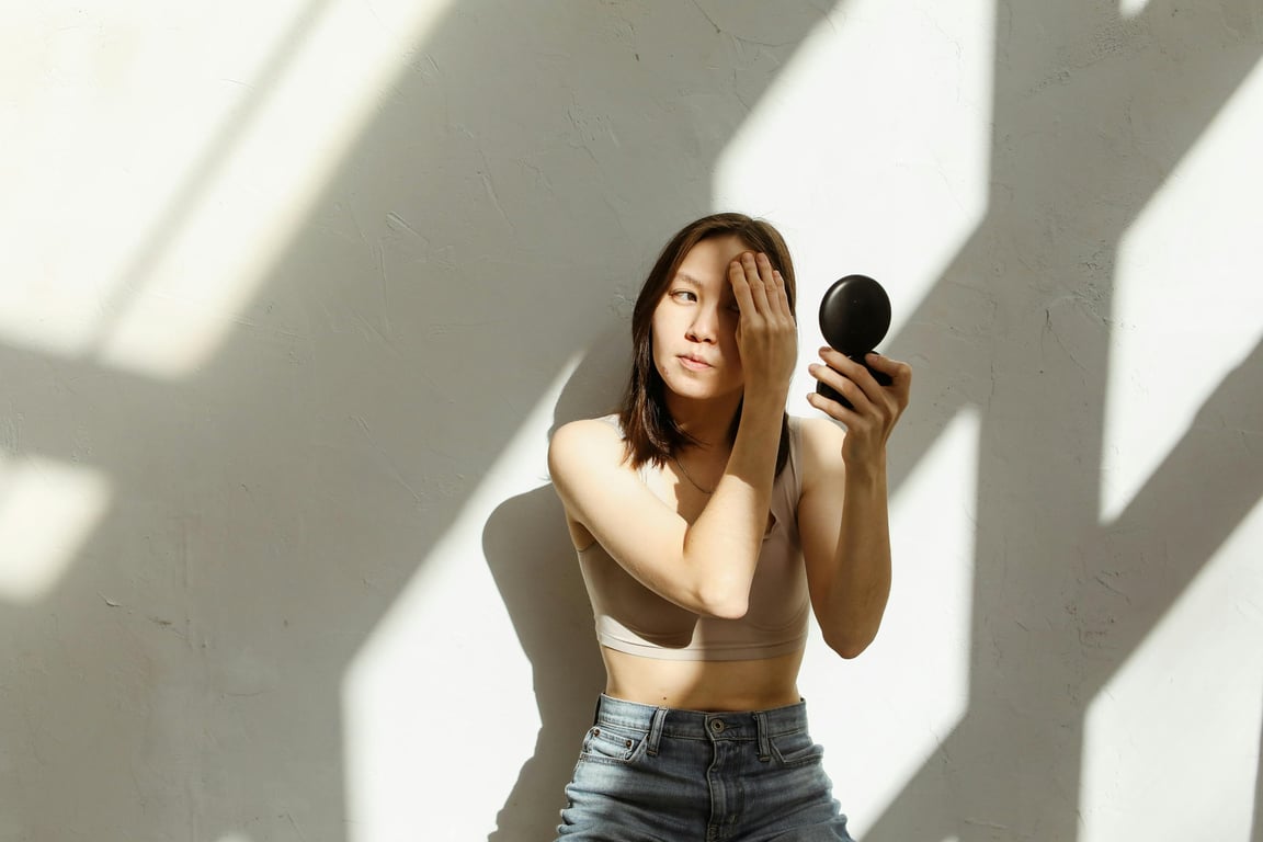 Young Asian woman in crop top applying makeup with face powder and mirror against a sunlit wall.