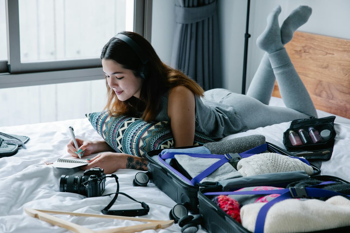 Woman lying on bed with headphones, writing in notebook, and packing suitcase.