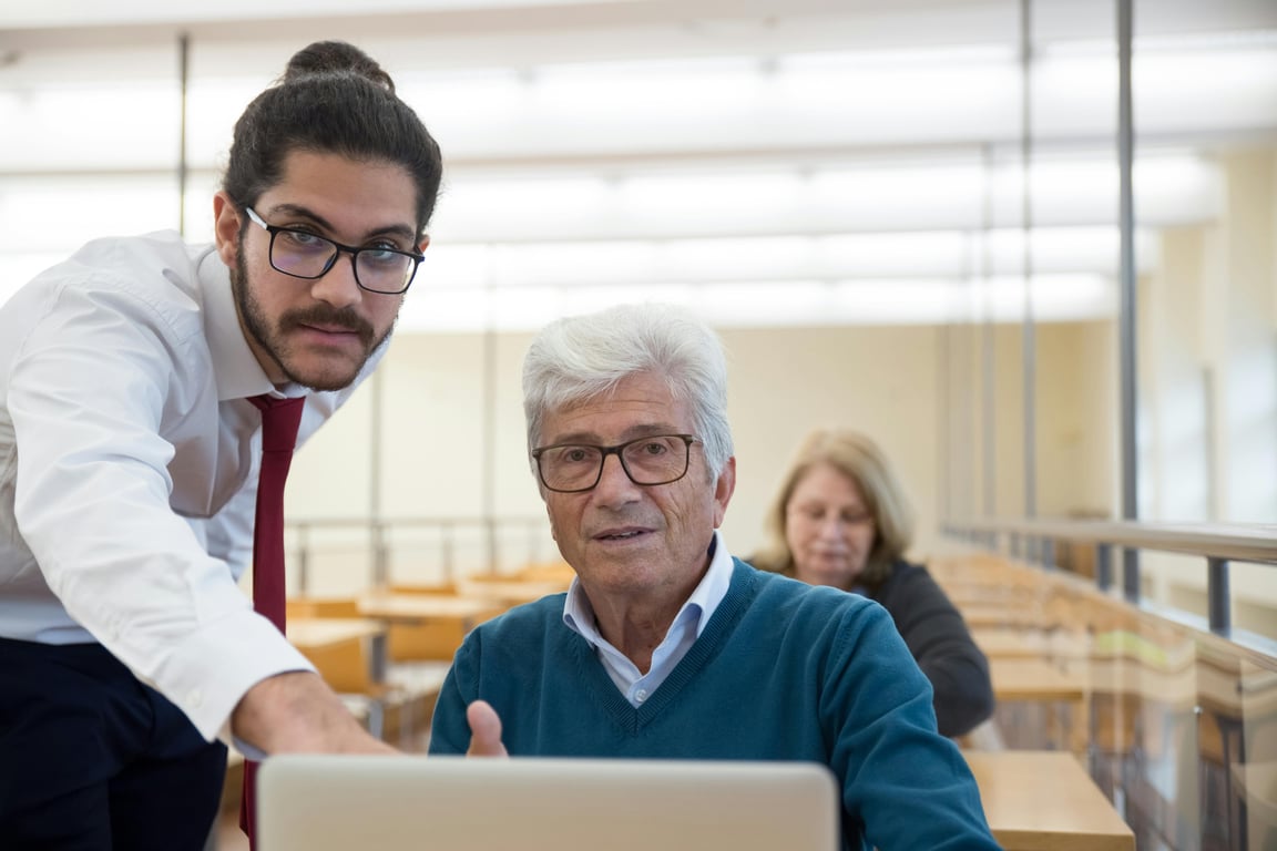 An elderly man learning computer skills with a teacher in a classroom setting, showcasing education through technology.