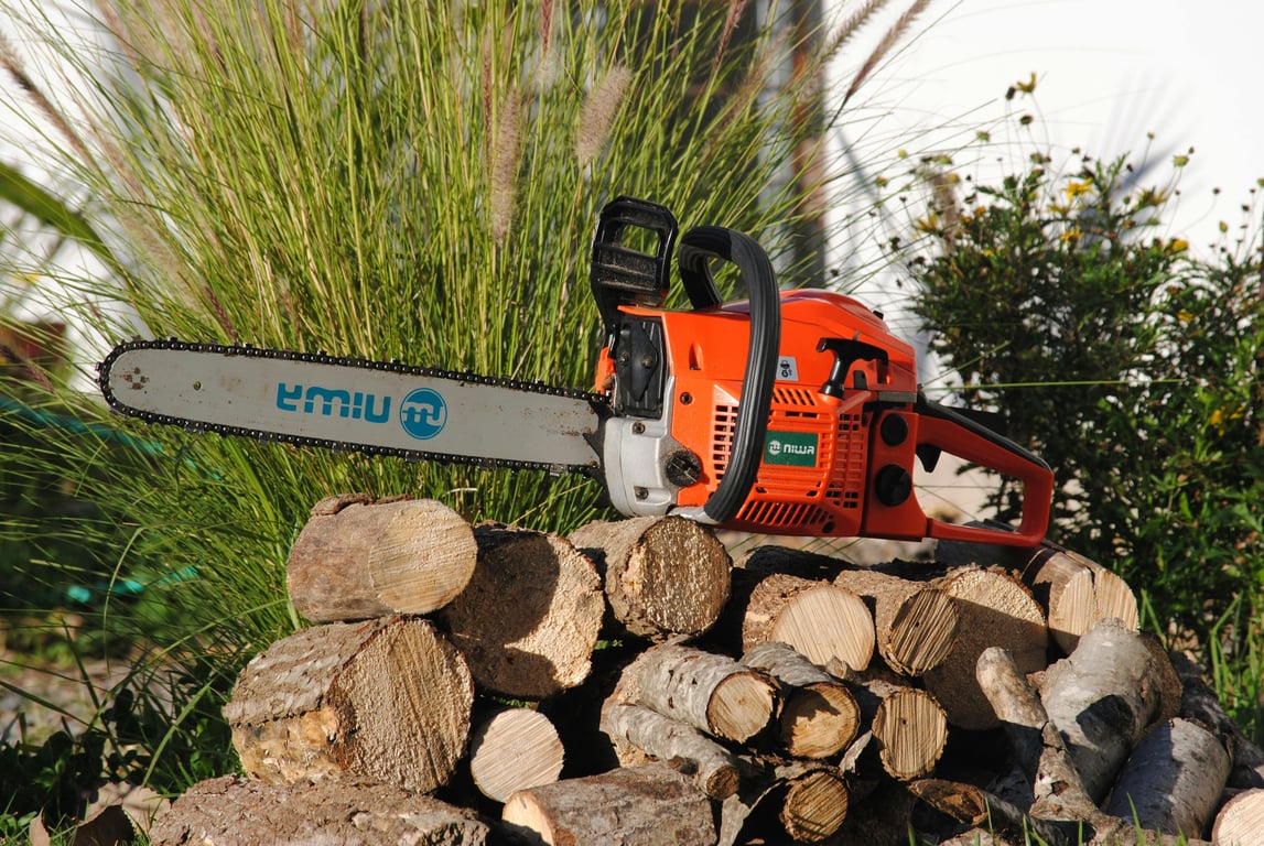 Bright orange chainsaw resting on a woodpile outdoors, surrounded by greenery.