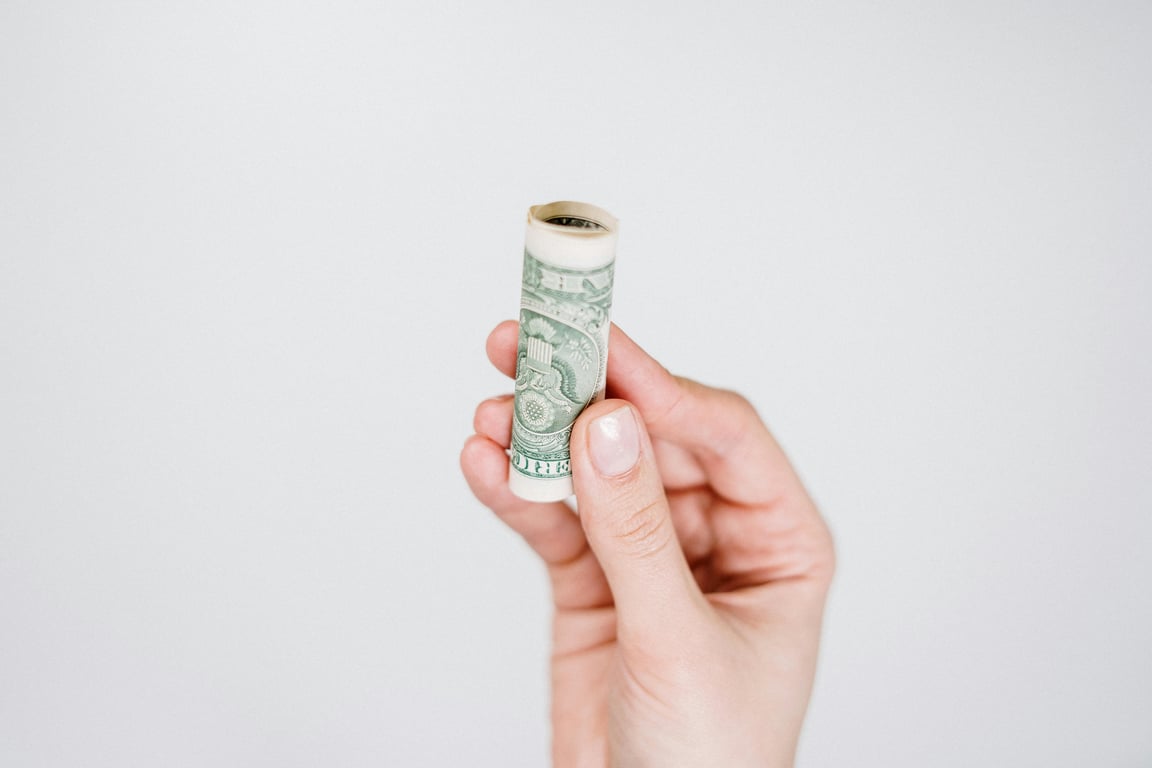 A hand holding a rolled US dollar bill against a plain background, symbolizing finance and savings.