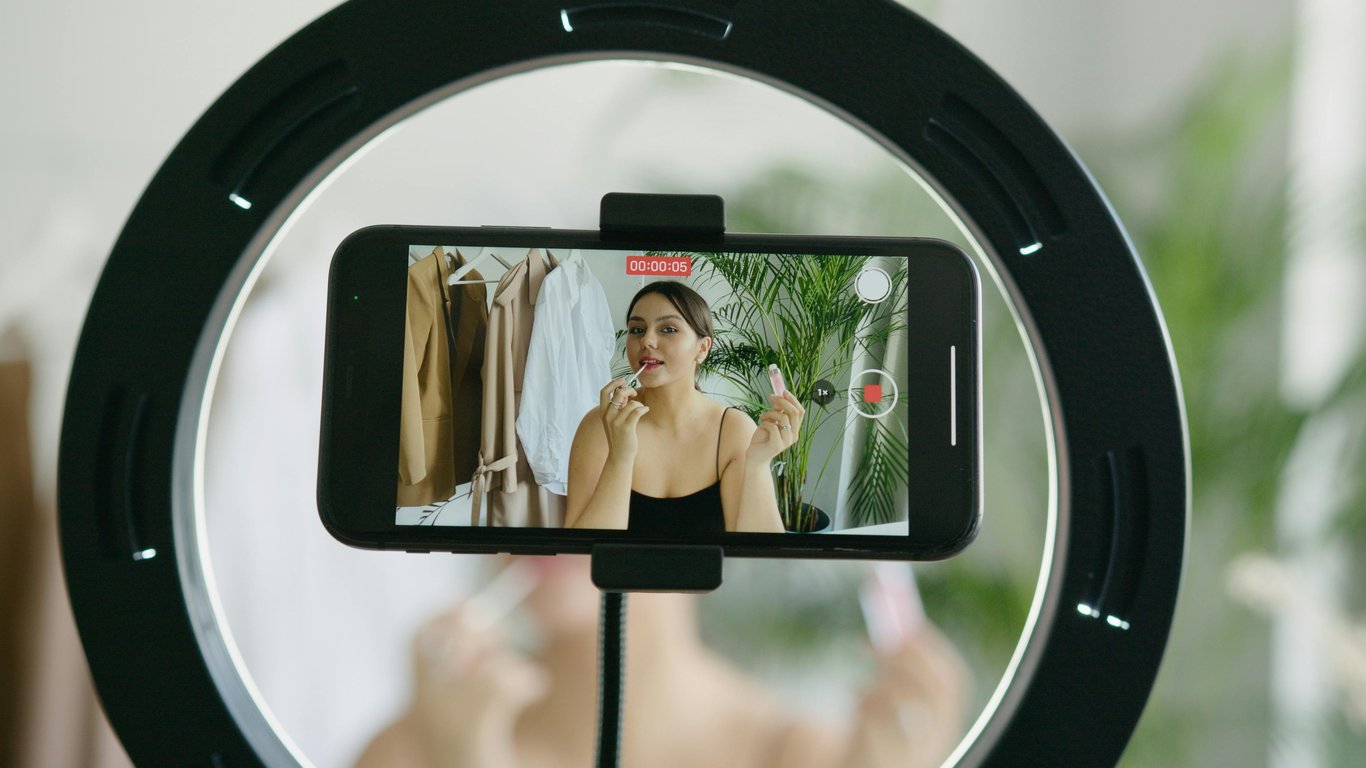 A beauty vlogger applying lipstick in a vlog recording using a smartphone and ring light.