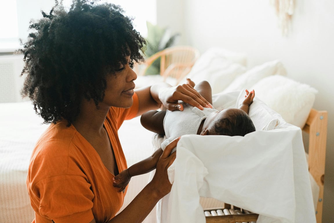 A mother lovingly looks at her baby lying on a bed in a warm, indoor setting.