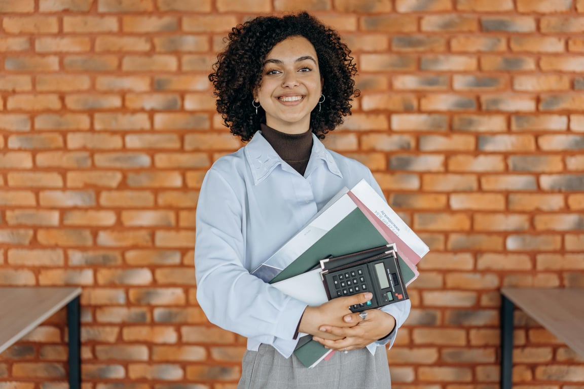 Cheerful woman with curly hair holding folders and a calculator against a brick wall.