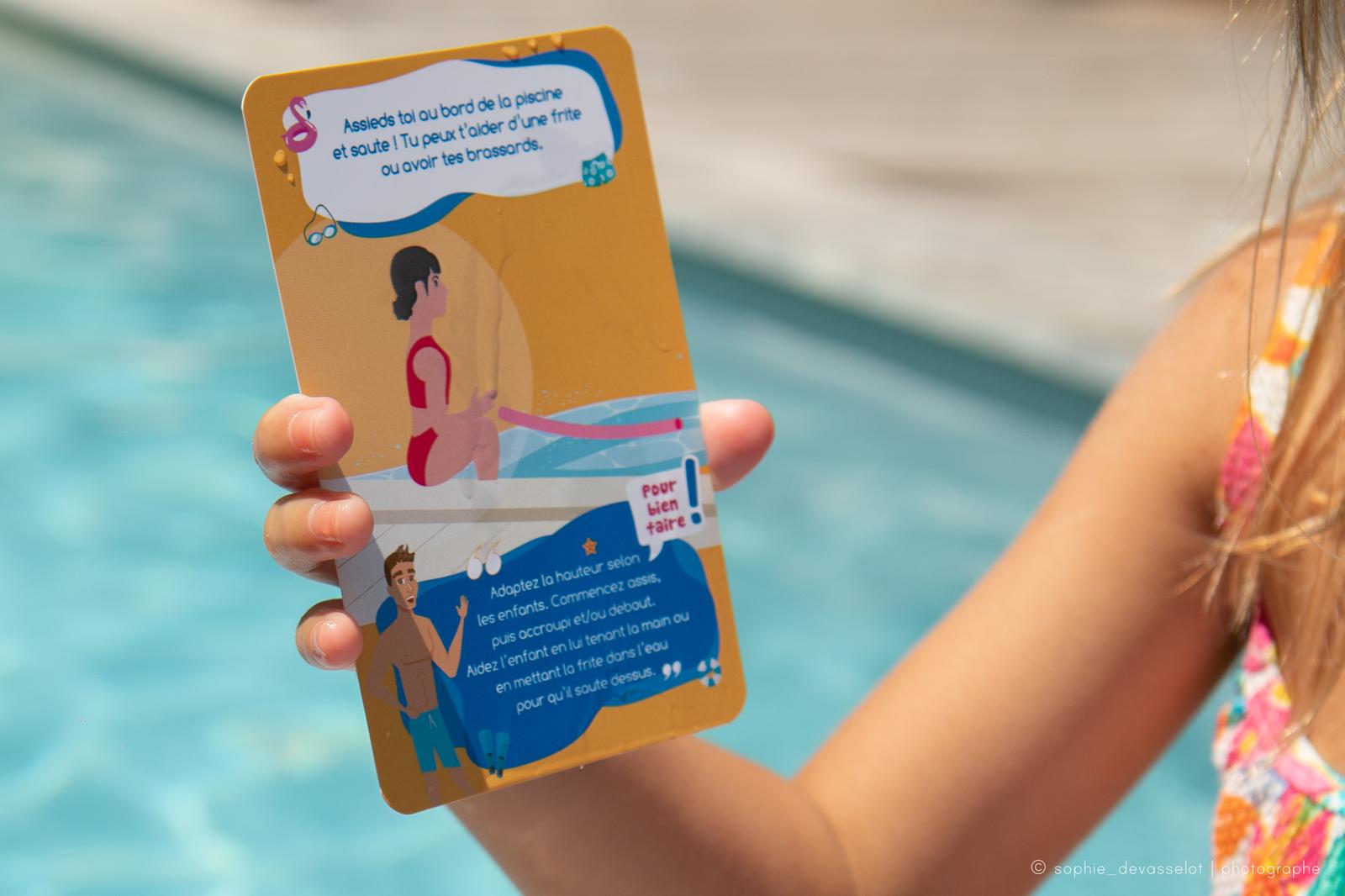 Une enfant tient une carte orange du jeu : la consigne « Assieds-toi au bord de la piscine et saute ! », l'illustration et la rubrique « Pour bien faire »