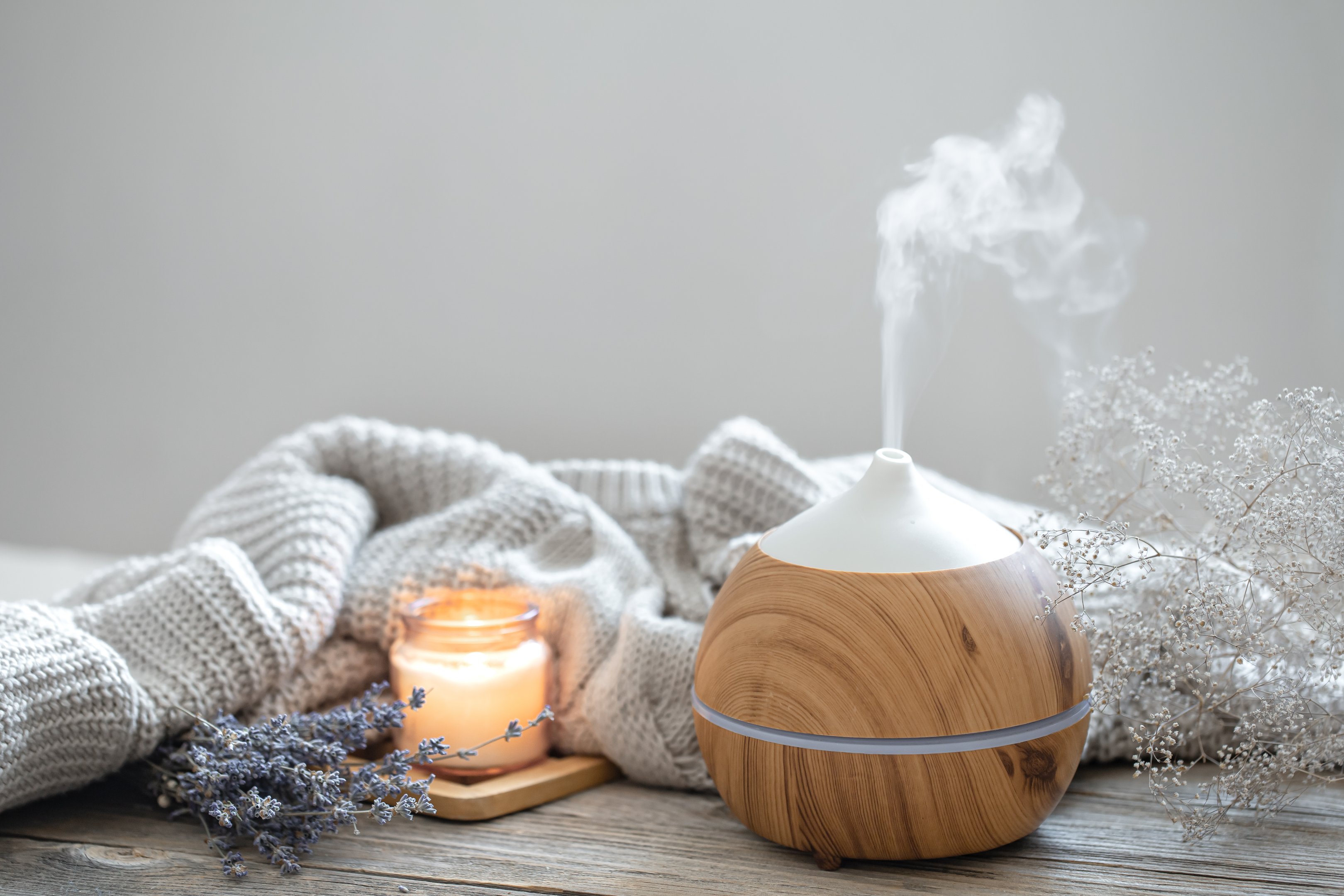 Wood-effect aroma diffuser releasing mist beside a candle and cosy knit blanket on a table