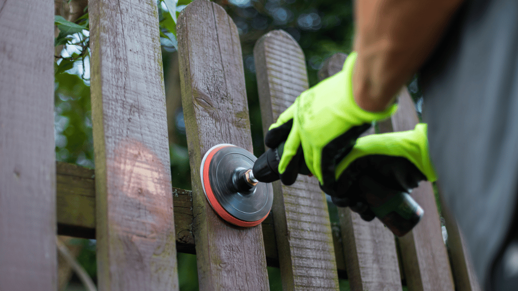 Person sanding a wooden garden fence with a power sander while wearing protective gloves.