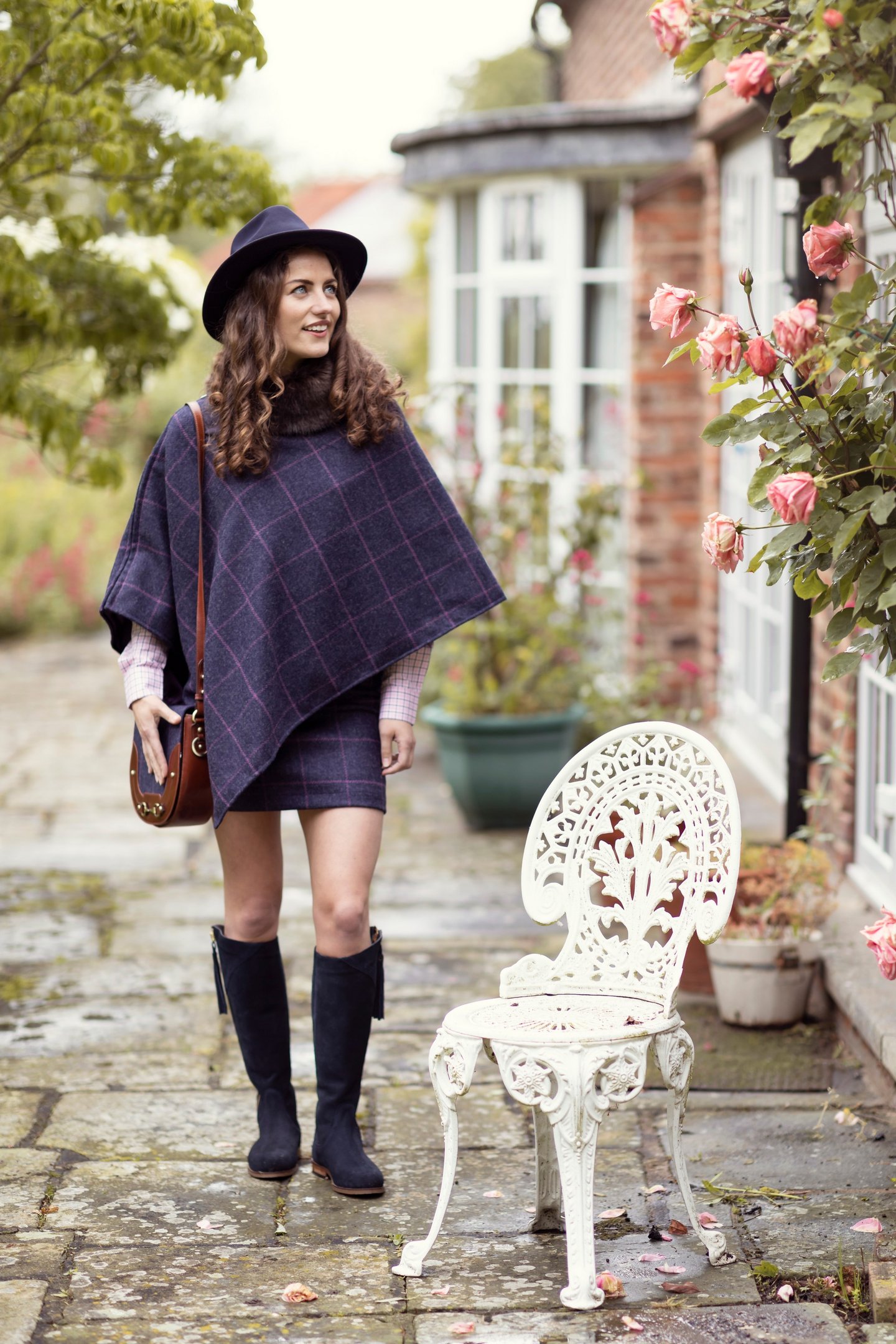 Frau in einem marineblauen karierten Tweedponcho und passendem Rock, dazu Filzhut und kniehohe Stiefel.