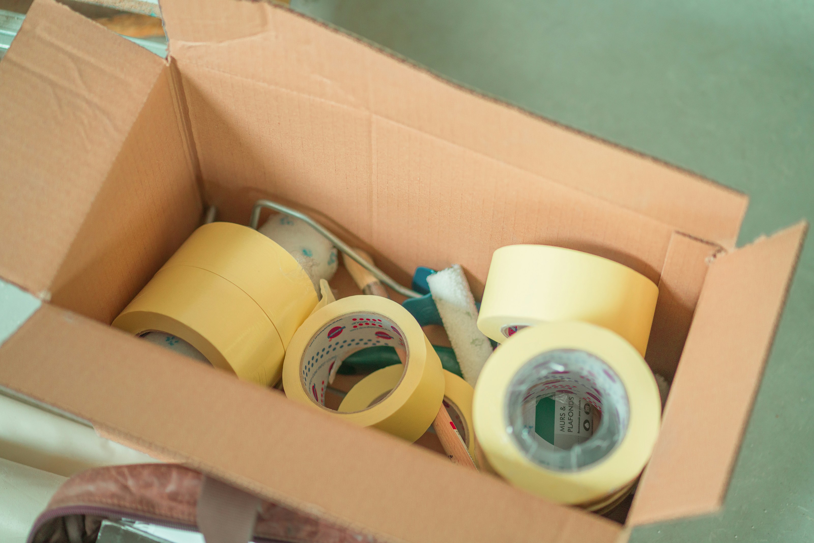Box of masking tape rolls and preparation tools used for protecting edges and surfaces before painting.