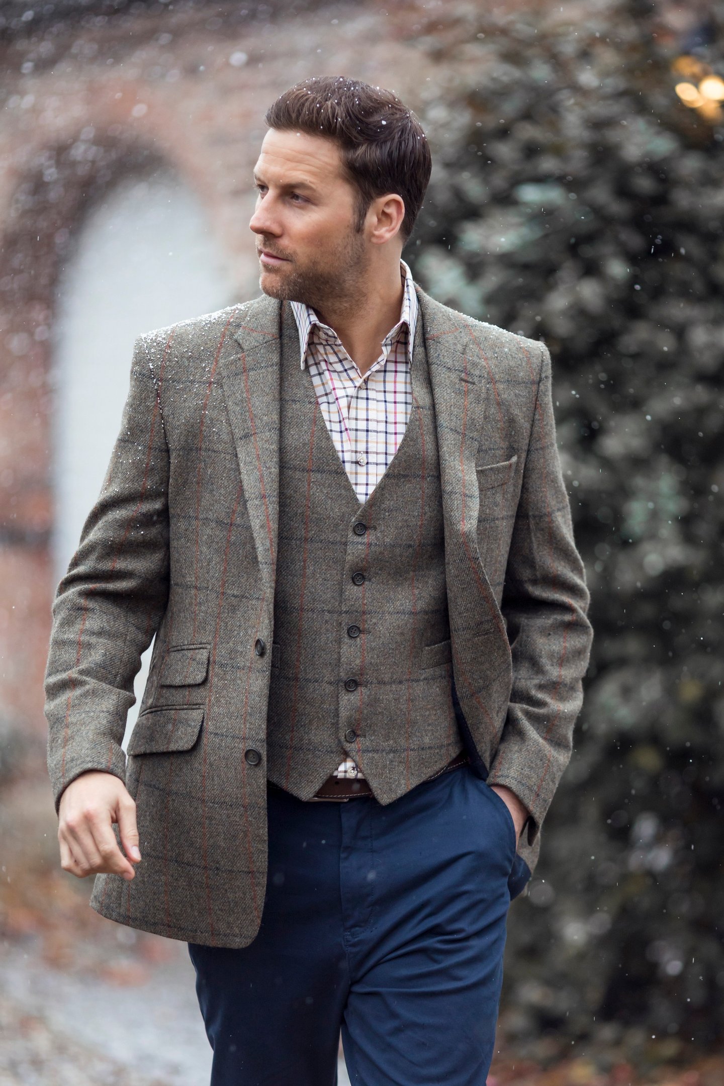 Ein Mann, der bei einer ländlichen Winterhochzeit eine braun karierte Tweedjacke und -weste mit marineblauer Hose trägt.