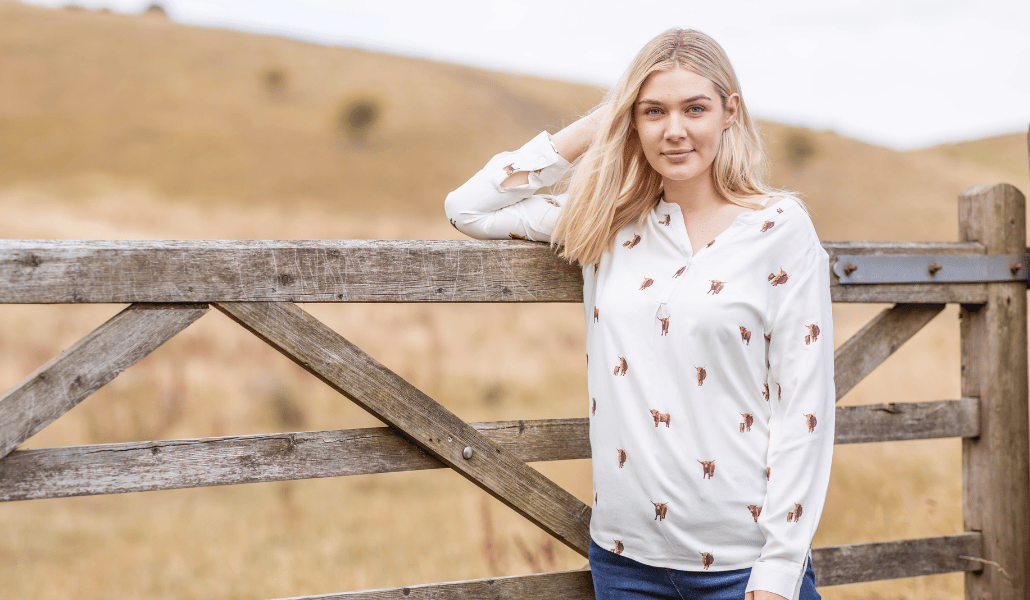 Frau in weißer Bluse mit Hochlandrindermuster und Jeans neben einem hölzernen Bauernhoftor