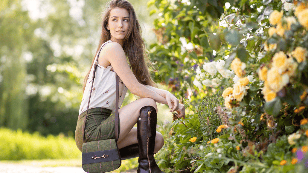 Woman wearing a printed sleeveless blouse, green tweed skirt and matching crossbody bag in a country garden.