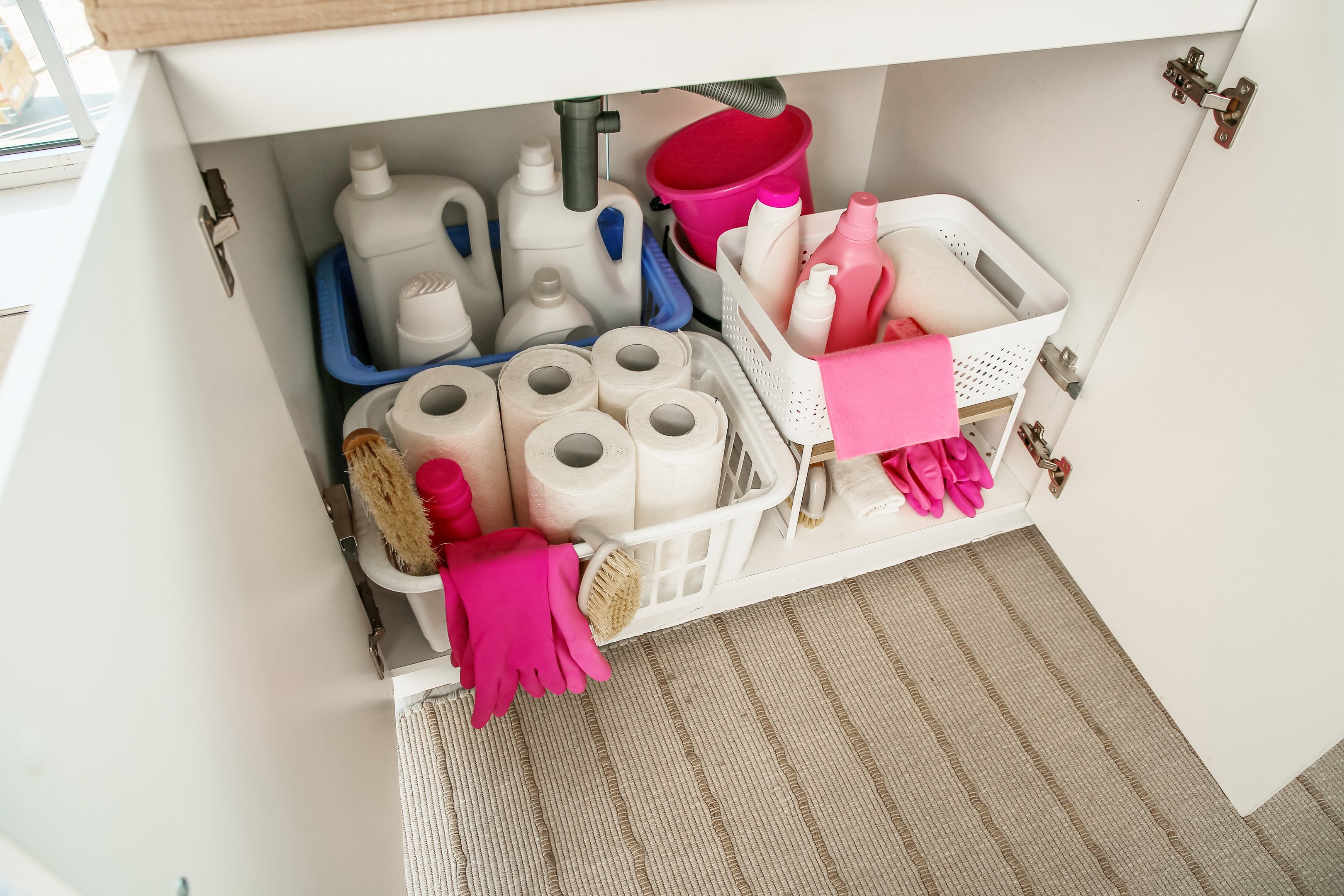 Under-sink cupboard with cleaning products, gloves and paper rolls stored neatly in pull-out baskets and caddies.