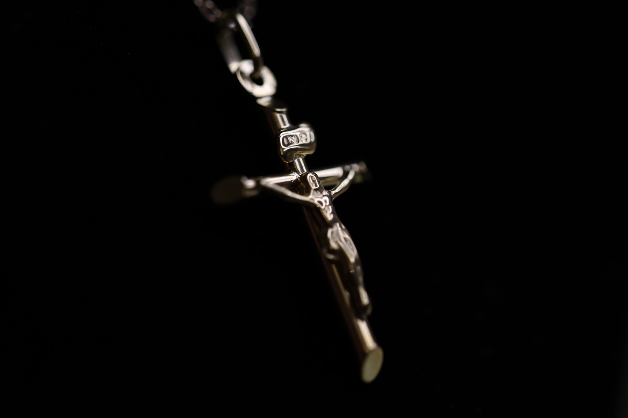 Polished silver crucifix pendant suspended on a chain against a black background, with a detailed corpus figure, INRI tablet at the top of the cross, and a small oval medallion on the left arm, lit with dramatic side lighting